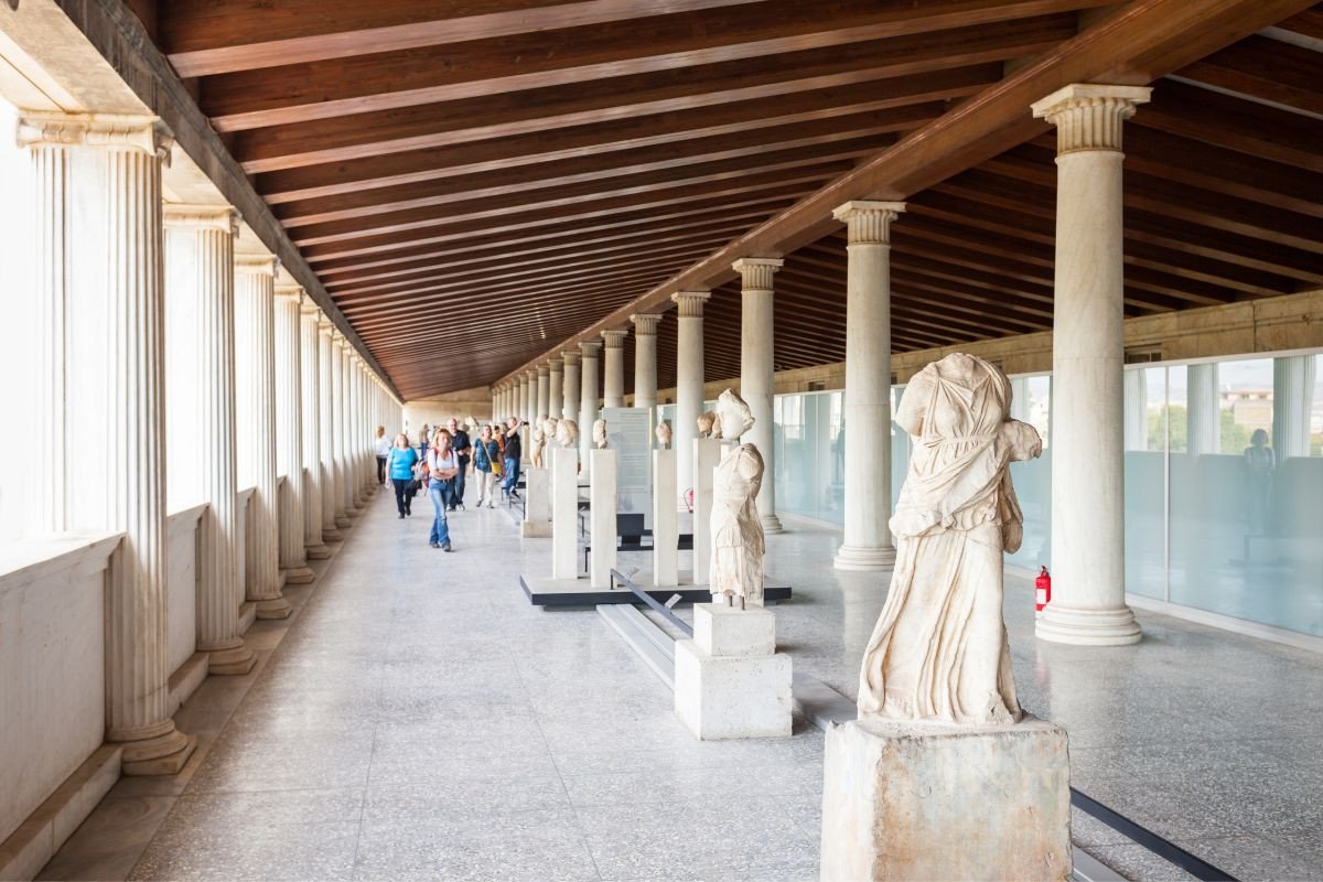 Visitors explore the Stoa of Attalos in the Ancient Agora of Athens, a covered walkway lined with tall, marble columns and statues of ancient figures. The ceiling features exposed wooden beams, and natural light floods the corridor from the left. The scene includes tourists walking and admiring the historical artifacts, creating a blend of ancient history and modern appreciation.